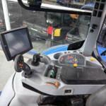 Interior of a blue tractor cab with a mounted touchscreen monitor, joystick controls, and a colored instrument panel; view through the windshield shows another vehicle nearby outside the building exterior.