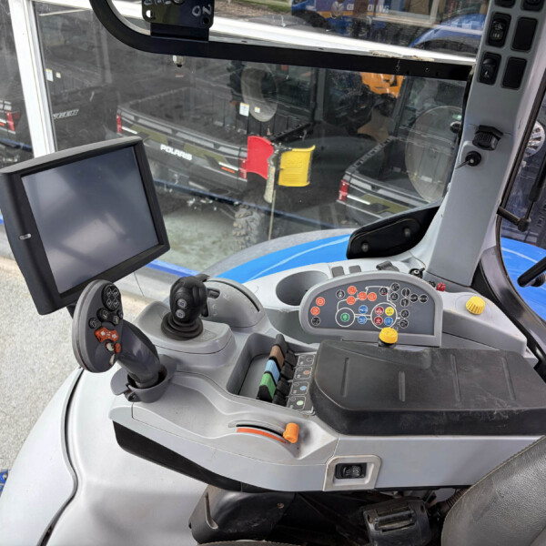 Interior of a blue tractor cab with a mounted touchscreen monitor, joystick controls, and a colored instrument panel; view through the windshield shows another vehicle nearby outside the building exterior.