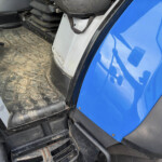 Interior of a blue vehicle cab showing a dirty rubber floor mat, pedals, and metal steps near the doorway.