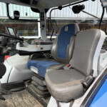 Interior cab of a heavy vehicle showing a blue and gray driver's seat, large steering wheel, and numerous control panels and levers in front of a dusty floor mat.