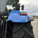 Close-up of a blue tractor front with exposed engine and oversized treaded tire in the foreground, at an industrial yard.