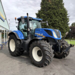 Blue New Holland tractor parked outside a dealership with a large tree in the background, Russells sign on the building nearby.
