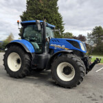 Blue New Holland tractor parked in an outdoor lot, large front tires visible and trees in the background.