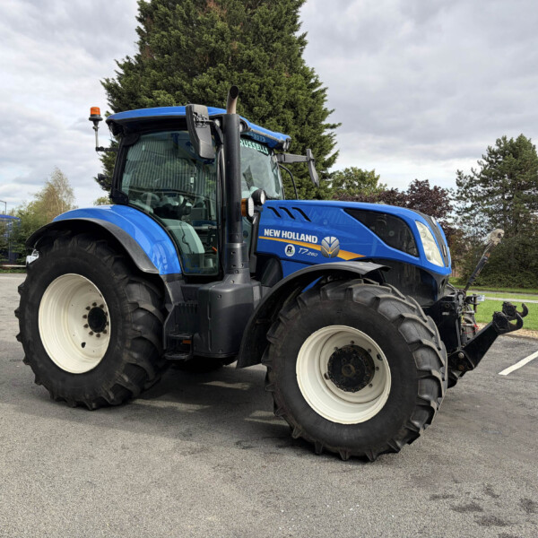 Blue New Holland tractor parked in an outdoor lot, large front tires visible and trees in the background.