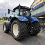 Blue New Holland agricultural tractor on display outside a dealership, front left view with oversized tires and orange warning lights on top of cab.
