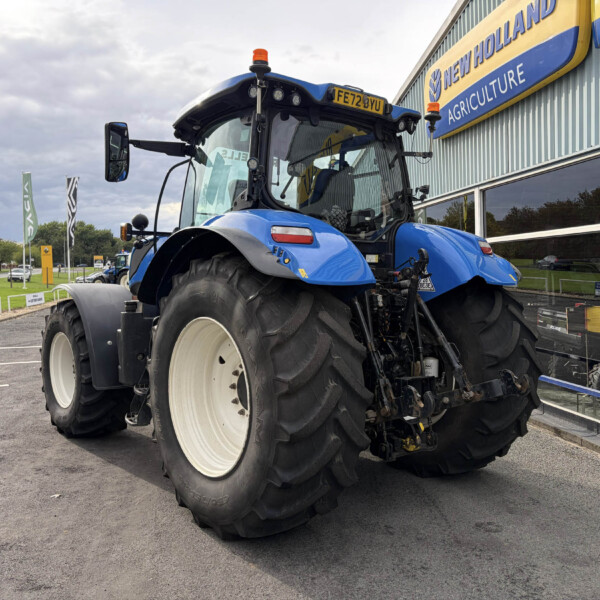 Blue New Holland agricultural tractor on display outside a dealership, front left view with oversized tires and orange warning lights on top of cab.