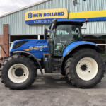 Blue New Holland agricultural tractor with large white-rimmed tires parked outside a blue-and-yellow New Holland sign on a corrugated metal building (agriculture showroom).