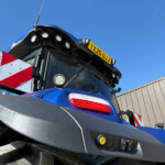 Close-up of a blue truck cab with roof lights, an orange beacon, and a yellow license plate reading YX26 UJJ against a clear blue sky.