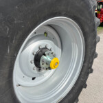 Close-up of a tractor wheel: large black tire with a gray rim, yellow hub, and lug bolts visible in the center. A red tractor is out of focus in the background.