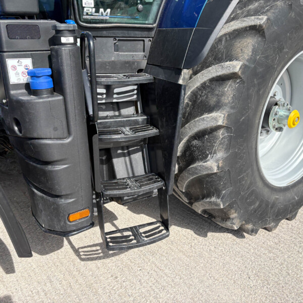Close-up of a tractor's step ladder and large treaded tire with a blue fuel cap nearby, showing rugged machinery details.