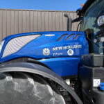 Blue New Holland tractor cab side with large front tire and fence background, lexicon branding visible on door area