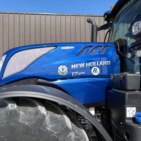 Blue New Holland tractor cab side with large front tire and fence background, lexicon branding visible on door area