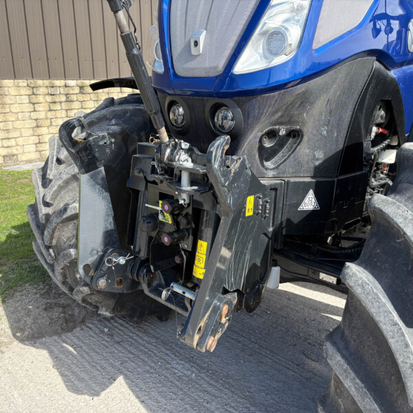 Close-up of a blue tractor's front end showing headlights and a damaged black bumper with exposed machinery and large treaded tire, parked near a brick wall and grass.