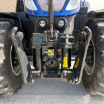 Front view of a blue tractor showing large muddy front tires, hitch and hydraulic assemblies with warning decals and two round headlights.