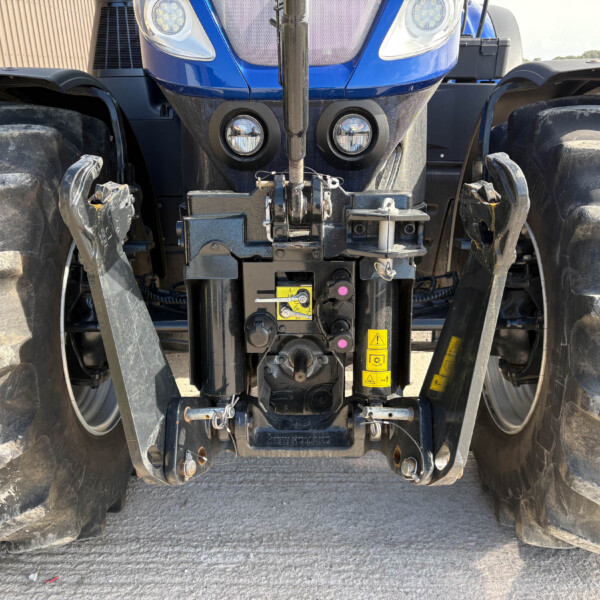 Front view of a blue tractor showing large muddy front tires, hitch and hydraulic assemblies with warning decals and two round headlights.