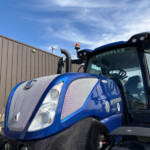 Blue tractor-trailer cab with orange beacon lights parked beside a wooden fence under a blue sky.