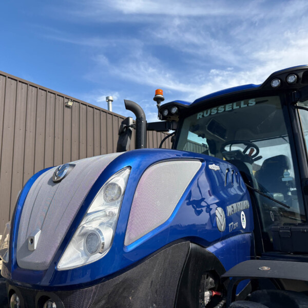 Blue tractor-trailer cab with orange beacon lights parked beside a wooden fence under a blue sky.