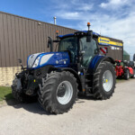 Blue New Holland tractor with oversized rear tires parked outside a brown corrugated metal building; other red tractors visible in the background.