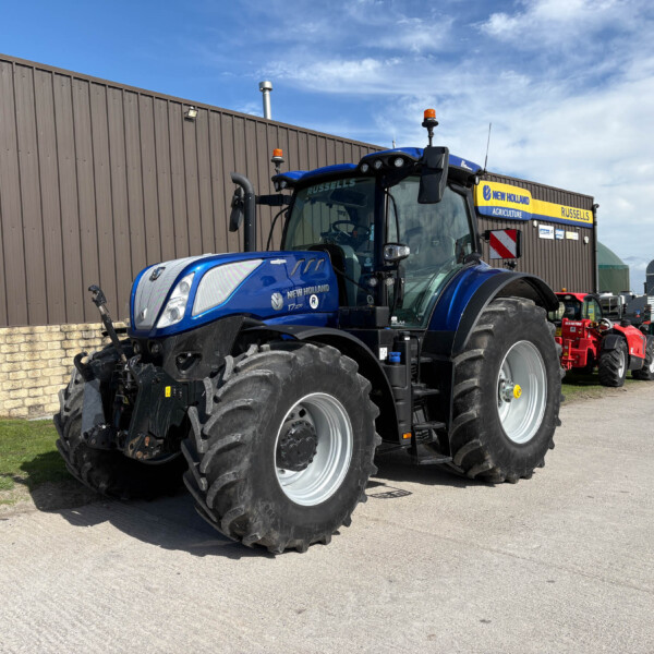 Blue New Holland tractor with oversized rear tires parked outside a brown corrugated metal building; other red tractors visible in the background.