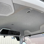 Interior view of a vehicle cab ceiling showing overhead speakers and a control panel near the windshield area.