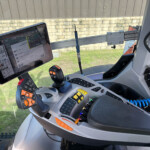Inside a tractor cab showing a mounted touchscreen display, multiple joysticks and control levers on a gray dashboard.
