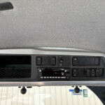 Truck cabin dashboard: overhead panel with multiple switches, a radio unit, and a grey fabric ceiling visible above the windshield area