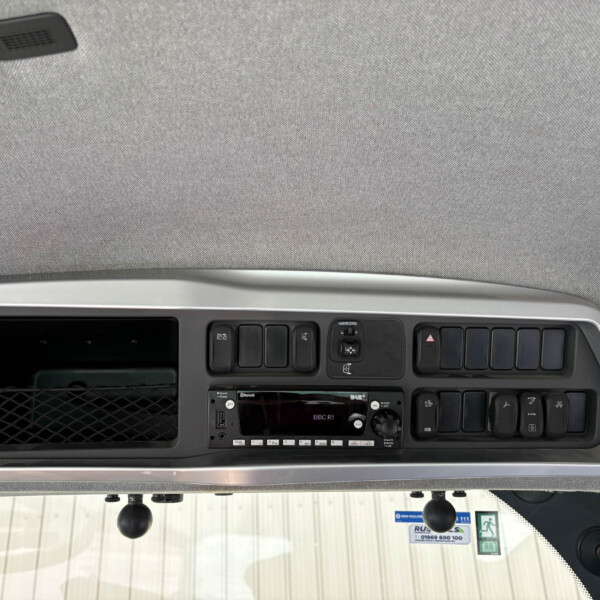 Truck cabin dashboard: overhead panel with multiple switches, a radio unit, and a grey fabric ceiling visible above the windshield area