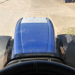 View from inside a blue truck cab looking down at the hood on a wide concrete lot, with open doors nearby and industrial buildings in the distance.