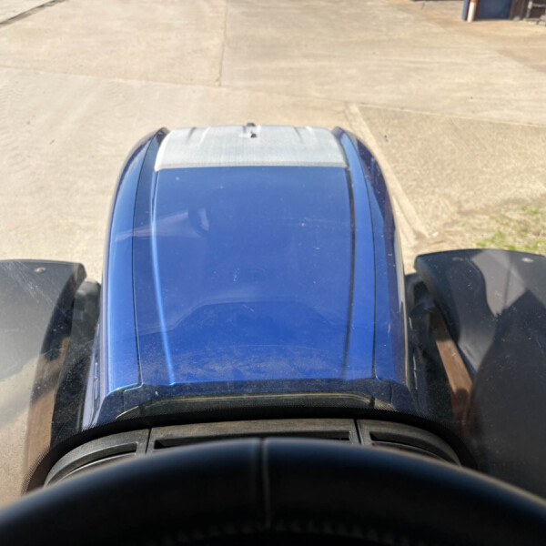 View from inside a blue truck cab looking down at the hood on a wide concrete lot, with open doors nearby and industrial buildings in the distance.