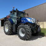Blue New Holland tractor with oversized tires parked outside a brown corrugated metal building under a clear blue sky.