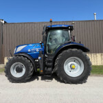 Blue New Holland tractor photographed from the front-left, with large tires and a brown industrial wall behind it.
