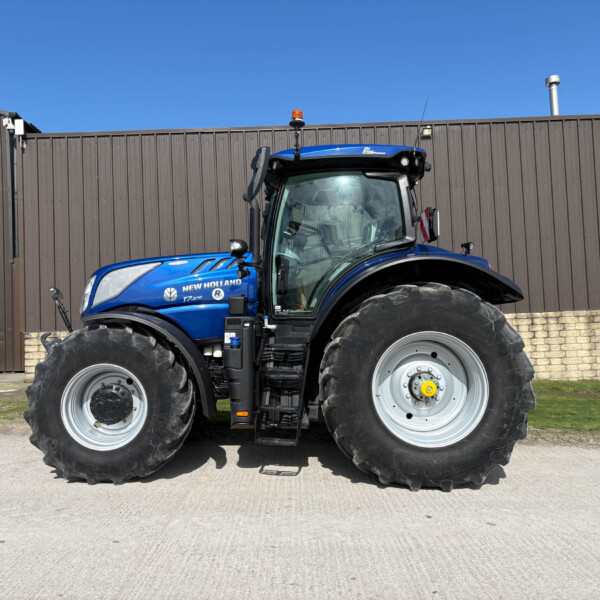Blue New Holland tractor photographed from the front-left, with large tires and a brown industrial wall behind it.