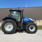 Front-side view of a blue New Holland tractor with oversized tires parked in front of a brown metal fence under a clear blue sky.