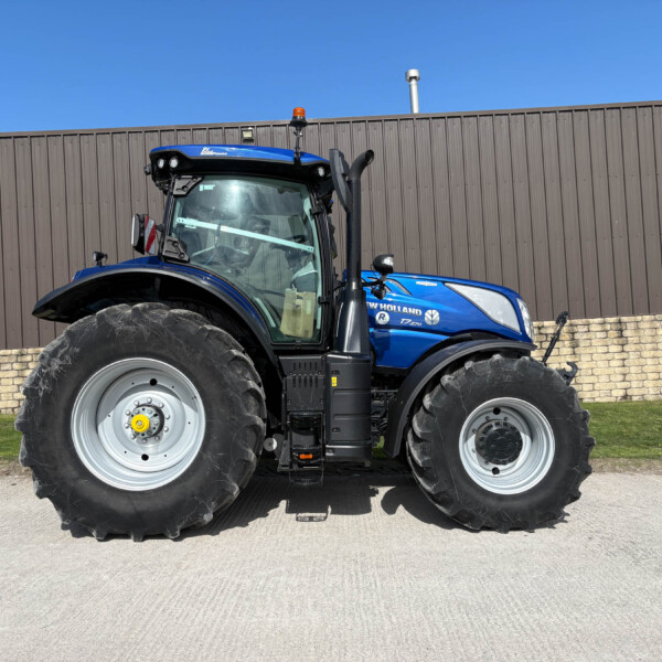 Front-side view of a blue New Holland tractor with oversized tires parked in front of a brown metal fence under a clear blue sky.