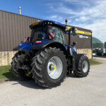 Blue agricultural tractor with oversized tires parked beside a brown corrugated metal wall on a sunny day.