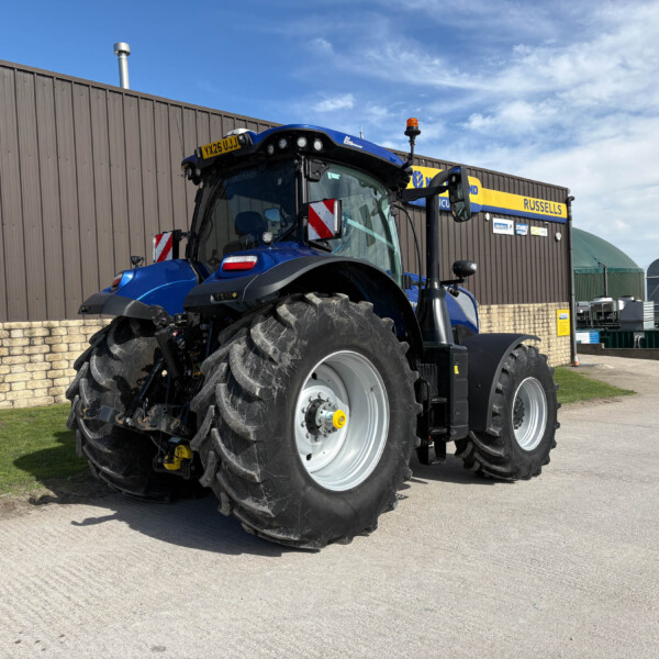 Blue agricultural tractor with oversized tires parked beside a brown corrugated metal wall on a sunny day.