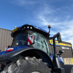 Front view of a blue New Holland tractor with oversized muddy front tires and an orange beacon on top.