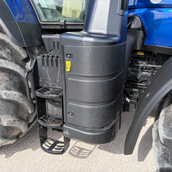Close-up of a blue tractor cab area with entry steps, a large front tire, and a black protective housing featuring a yellow caution sticker.