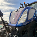 Close-up of a blue tractor front with a chrome grille, headlights, and an orange beacon on top against a blue sky.