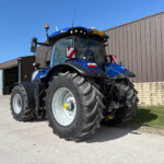 Blue New Holland tractor parked outside a brown metal industrial building on a sunny day, front view visible with large tires.