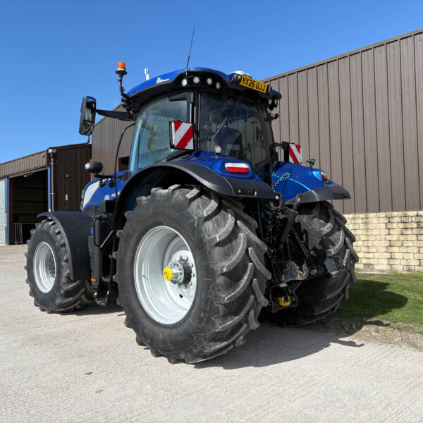 Blue New Holland tractor parked outside a brown metal industrial building on a sunny day, front view visible with large tires.