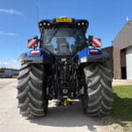 Rear view of a blue agricultural tractor with oversized rear tires in a yard beside a brown metal building under a clear blue sky.