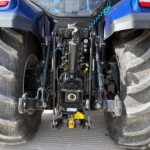 Blue tractor rear with exposed hydraulic valves and yellow three-point hitch between large muddy rear tires