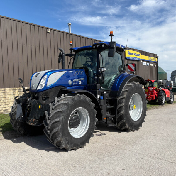 Blue New Holland tractor in front of a brown metal building with an orange beacon and other farming gear in the background.