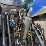 Close-up of a tractor front-end showing hydraulic components, hoses, and blue coiled air lines amid dusty machinery.