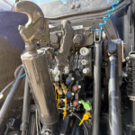 Close-up of a heavy machinery cockpit showing hydraulic levers, cables, and metal control blocks with a blue spiral air hose nearby.