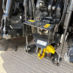 Close-up of heavy machinery undercarriage showing hydraulic components, metal arms, wiring, and yellow safety hooks with warning labels
