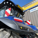 Low-angle close-up of a blue tractor, showing a large muddy front tire, lights, and a yellow license plate against a wooden building backdrop.