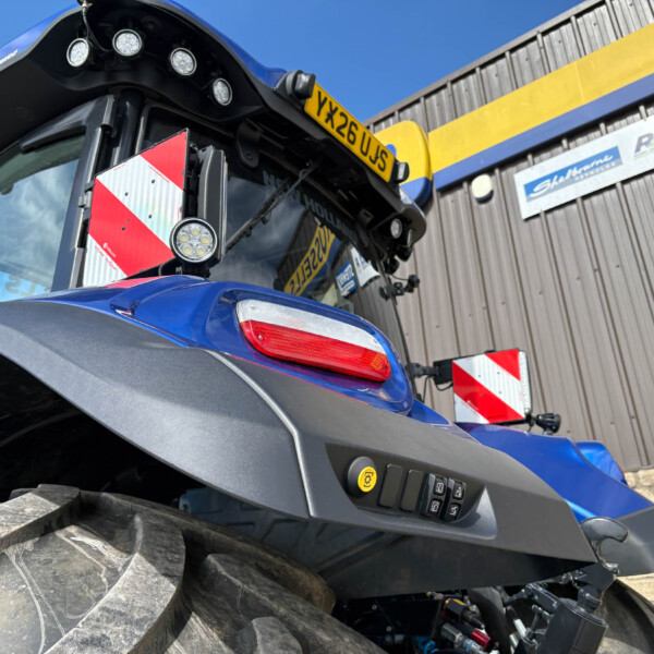 Low-angle close-up of a blue tractor, showing a large muddy front tire, lights, and a yellow license plate against a wooden building backdrop.