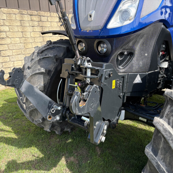 Blue tractor front end with hydraulic hitch and large tires, parked on grass beside a brick wall.